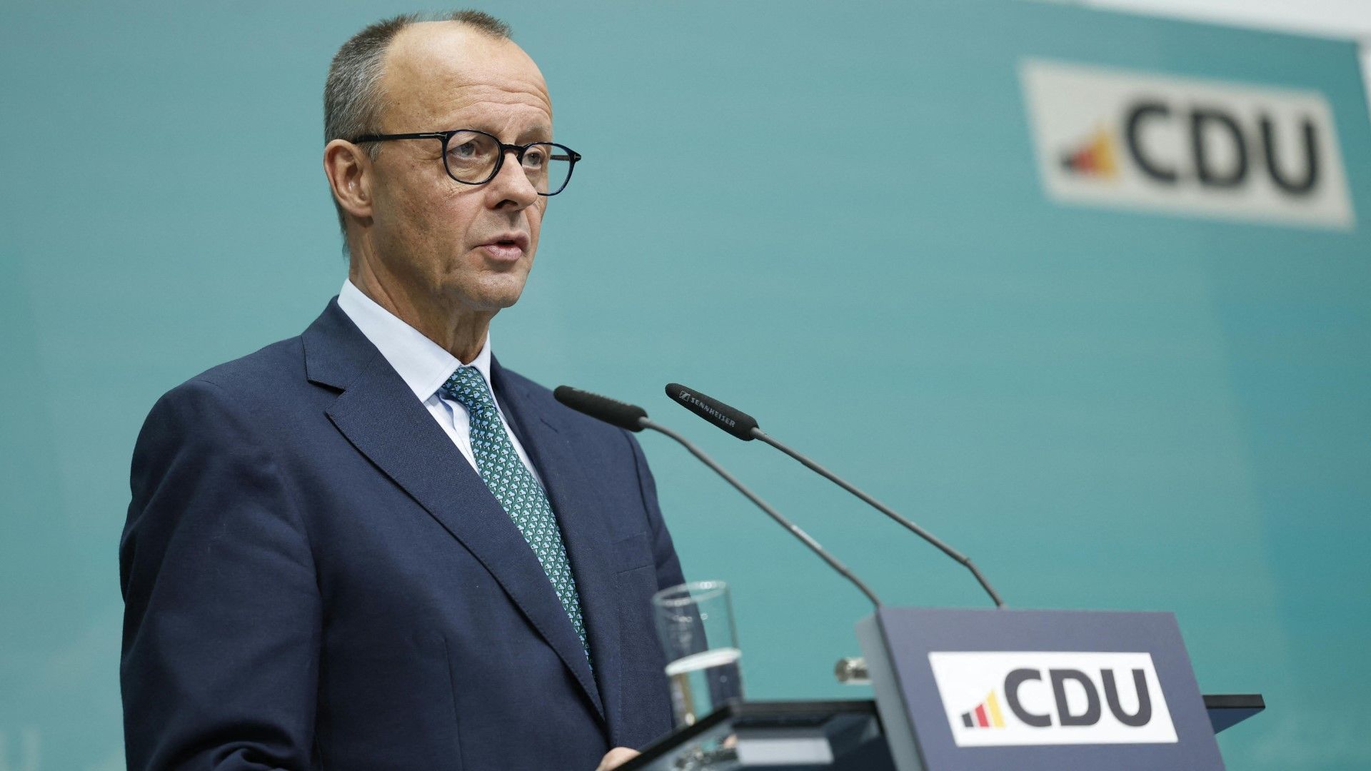 Friedrich Merz in einem dunklen Anzug mit weißem Hemd und grüner Krawatte steht an einem Rednerpult mit dem Logo der CDU. Er trägt eine Brille und spricht in ein Mikrofon. Im Hintergrund ist eine türkisfarbene Wand mit dem CDU-Logo zu sehen.
