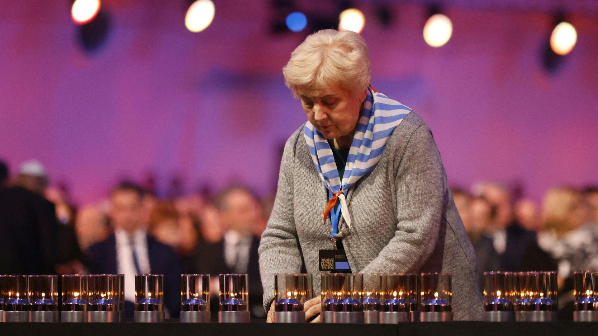 Eine ältere Frau in einem grauen Mantel und einem gestreiften Halstuch lehnt sich über mehrere Gedenkkerzen bei einer Zeremonie in Auschwitz. Im unscharfen Hintergrund sind weitere Teilnehmende zu erkennen.