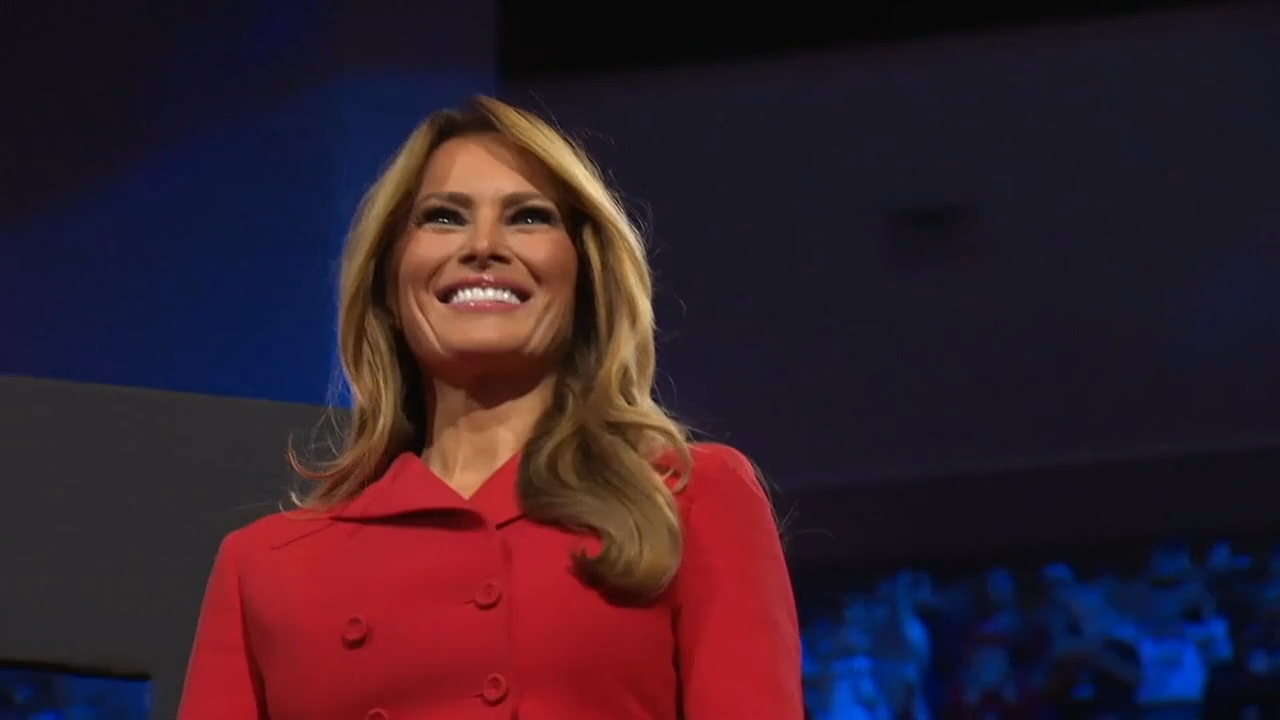 Melania Trump lächelnd. Sie hat einen roten Blazer an. Melania Trump lächelnd. Sie hat einen roten Blazer an.